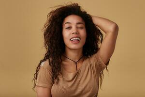 Indoor photo of young lovely curly dark skinned brunette female with natural makeup keeping one eyes closed while looking cheerfully at camera, posing over beige background