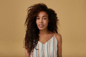 Beautiful young long haired curly dark skinned female with natural makeup looking pensively at camera while standing over beige background with hands down photo