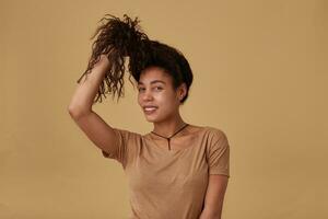 Positive young lovely brown haired curly female with dark pulling her hair with raised hand and smiling cheerfully at camera, isolated over beige background photo