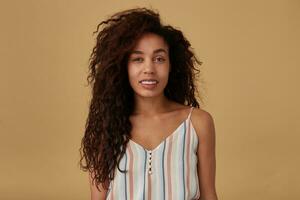 Studio shot of young charming brown haired curly female with dark skin keeping her hands down while posing over beige background, being in nice mood and smiling pleasantly photo