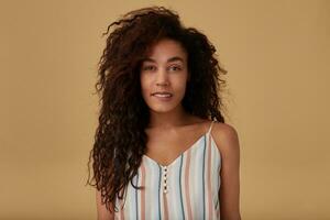 Flirty young pretty curly dark skinned brunette lady with casual hairstyle biting underlip while looking gently at camera, isolated over beige background in striped top photo