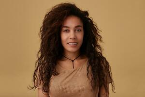 Portrait of young charming brown haired curly dark skinned lady with casual hairstyle looking at camera with charming smile while standing over beige background photo