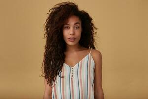 Horizontal photo of young attractive dark skinned woman with brown long curly loose hair looking attentively at camera while standing over beige background