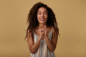 Positive young attractive curly dark skinned brunette woman in summer striped blouse keeping raised hands folded and giving glagly wink at camera, isolated over beige background photo