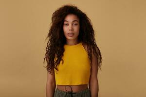 Studio photo of pretty curly brunette dark skinned woman with natural makeup looking at camera with calm face while posing over beige background with hands down