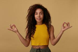 estudio foto de joven atractivo Rizado morena hembra con natural maquillaje formando dedos en namaste firmar y mirando tranquilamente a cámara, aislado terminado beige antecedentes