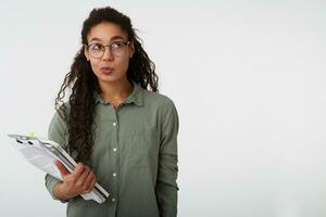 Puzzled young pretty dark skinned curly female with casual hairstyle pusing her lips while looking pensively upwards and keeping textbooks in raised hand, isolated over white background photo