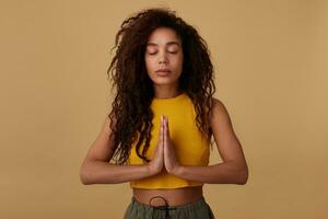 Pleasant looking young calm curly brunette dark skinned woman keeping her eyes closed while meditating and raising folded hands, isolated over beige background photo