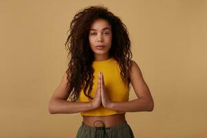 Studio photo of young pretty long haired curly brunette lady with dark skin keeping raised palms together while standing over beige background in yellow top