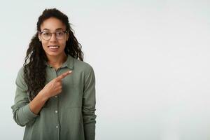 Cheerful young lovely brown-eyed curly dark skinned woman in glasses looking at camera with charming smile while pointing aside with index finger, posing over white background photo