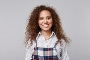 Portrait of glad young attractive brown haired curly lady being in nice mood and smiling cheerfully at camera, isolated over grey background in elegant wear photo