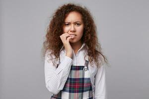 Indoor shot of confused young curly brunette female biting worringly nails and frowning eyebrows while looking at camera, standing over grey background photo