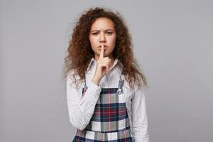 Severe young long haired curly brunette woman frowning eyebrows and keeping forefinger on her lips while asking to keep silence, isolated over grey background photo