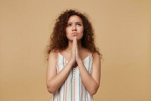 Serious young attractive curly brunette lady with casual hairstyle folding hands in praying gesture and looking sadly upwards, isolated over beige background photo