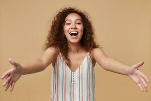 Portrait of young pretty cheerful brown haired curly female keeping her hands wide opened while looking joyfully at camera, isolated over beige background photo