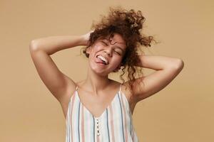 Funny shot of young pretty curly brunette woman holding her hair with raised hands and sticking happily out tongue, standing over beige background in casual wear photo