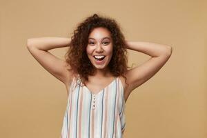 Studio photo of young lovely curly brunette woman with casual hairstyle keeping raised hands on her head while looking cheerfully at camera, isolated over beige background