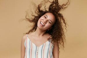 Indoor photo of cheerful young attractive woman waving her curly brown hair while looking happily at camera with broad smile, posing over beige background