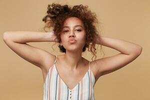 Portrait of positive young brown haired curly female keeping raised hands on her head and pursing lips while looking at camera, isolated over beige background photo