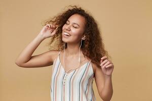 Indoor photo of young cheerful brown haired curly female with casual hairstyle dancing with raised hands and smiling gladly, standing over beige background