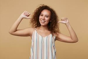 Studio photo of young joyful curly brunette woman with casual hairstyle pointing on herself with raised hands and smiling broadly, posing over beige background