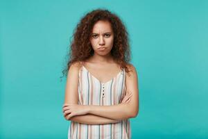 Indoor photo of severe young curly brunette female folsing her hands on chest while looking seriously at camera, isolated over blue background in summer blouse
