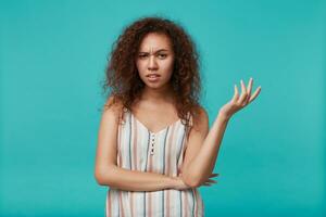 Puzzled young brown-eyed curly brunette lady frowning her eyebrows and raising confusedly hand while looking at camera, isolated over blue background photo