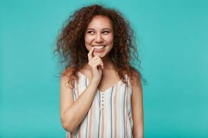 Indoor photo of young pretty brown haired curly lady with natural makeup smiling cheerfully while looking dreamily aside and keeping forefinger on her mouth, isolated over blue background