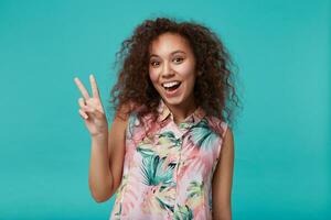 Joyful young lovely curly brunette female forming with raised fingers victory sign and looking happily at camera with wide smile, isolated over blue background photo