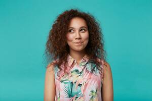 Portrait of charming young curly brunette woman with natural makeup biting her lips while looking positively aside, isolated over blue background with hands down photo