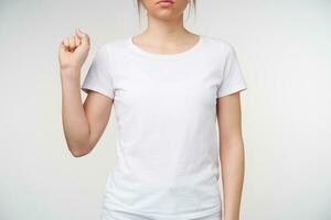 Studio photo of young female with nude manicure clenching fist while showing letter s on sign language, being isolated over white background in white t-shirt