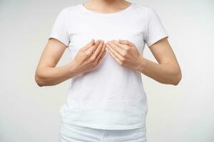 Studio photo of young female dressed in white t-shirt learning sign language while standing over white background, keeping palms in front of her chest while showing word book