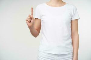 Studio photo of young woman dressed in white t-shirt while posing over white background, keeping forefinger raised while counting one. Hands and gesturing concept