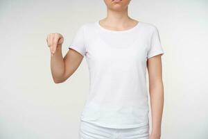 Cropped shot of young woman dressed in white t-shirt showing down with two fingers while learning deaf language, being isolated over white background photo