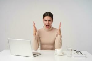 Irritated young attractive dark haired lady raising emotionally hands while shouting angrily with wide mouth opened, dressed in beige poloneck while sitting over white background photo