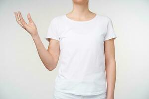 Cropped shot of young lady dressed in white t-shirt raising her palm while expressing thoughts using deaf language, standing over white background photo