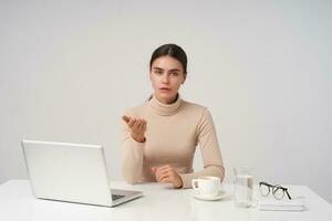 Indoor photo of young pretty dark haired lady dressed in beige poloneck sitting at table with laptop and cup of coffee, raising perplexedly palm while looking at camera with puzzled face
