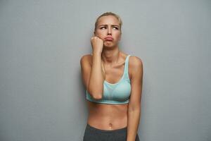 Frowning sad young slim blonde woman looking aside and pouting her lips, leaning head on raised hand while standing over grey background, dressed in sporty wear photo