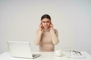 Stressed young dark haired woman with ponytail hairstyle keeping index fingers on temples and frowning her face, having strong headache while sitting over white background photo