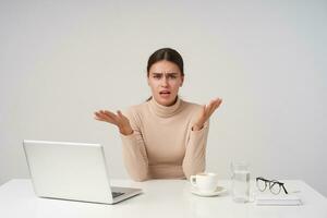 Agitated young beautiful dark haired female raising hands while looking confusedly at camera, working at office with modern laptop, wearing formal clothes while posing over white background photo