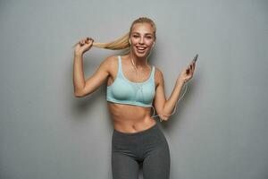 Joyful beautiful sporty blue-eyed lady with blonde hair looking at camera happily with broad smile, holding smartphone in raised hand and wearing earphones, posing against grey studio background photo