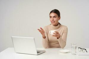 Beautiful young dark haired business lady having meeting with colleagues, holding cup of tea while sitting over white background in beige poloneck and keeping her hand raised photo