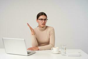 Puzzled young pretty brunette lady with ponytail hairstyle frowning eyebrows and raising confusedly her palm while looking aside, posing over white background photo