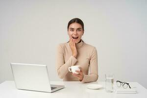 Excited young lovely brunette woman holding hand on her cheek and looking at camera with wide eyes and mouth opened, holding cup of tea while posing over white background photo