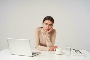 Bored young attractive brunette female leaning her head on raised hand while sitting at table, working with laptop at office, keeping her lips folded while looking at camera photo