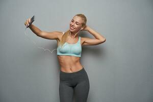 Good looking young cheerful slim female with long blonde hair making photo of herself with smartphone while posing over grey background, wearing mint sporty top and grey leggins