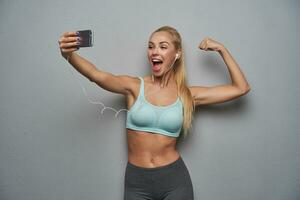 Happy pretty sporty blonde lady with ponytail hairstyle keeping mobile phone in raised hand and demonstrating her strong biceps, standing over grey background with wide joyful smile photo