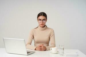 Serious young dark haired businesswoman wearing eyewear and formal clothes while sitting at table with laptop, folding hands on countertop while posing over white background photo