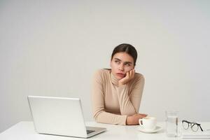 Weary young attractive dark haired lady leaning her chin on raised hand and looking dreamily aside, having long working day and counting time, isolated over white background photo