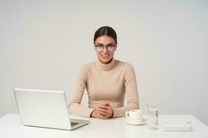 Shot of young cheerful beautiful brunette lady having meeting at office, being in nice mood and smiling pleasantly, holding hands on table while sitting over white background photo